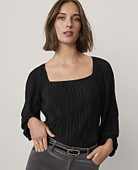 A woman wearing a black pleated top and grey trousers stands against a plain light grey background. carousel Product Image 1