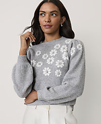 A woman wearing a grey floral-patterned sweater and white trousers poses against a plain light-colored background. carousel Product Image 1