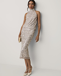 A woman wearing a shimmering champagne-colored sequin midi dress and heels stands against a plain white wall. carousel Product Image 1