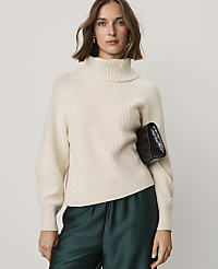 A woman wearing a cream-colored turtleneck sweater and dark green satin pants holds a black clutch against a plain light gray background. carousel Product Image 1