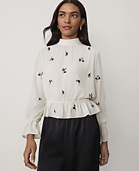 A woman wearing a white floral-embroidered blouse and black trousers stands against a plain light-colored background. carousel Product Image 1