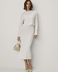 A woman wearing a white ribbed knit two-piece set and holding a gold handbag stands against a plain white wall. carousel Product Image 1
