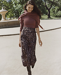 A smiling woman wearing a mauve top and a paisley skirt walks along a paved path with lush green trees in the background. carousel Product Image 4