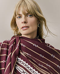 A blonde woman wearing a patterned burgundy and beige scarf and gold earrings poses against a plain, neutral background. carousel Product Image 4