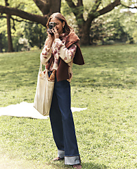 A woman in a patterned sweater and wide-leg jeans takes a photo with a vintage camera while standing on a grassy lawn with large trees in the background. carousel Product Image 5