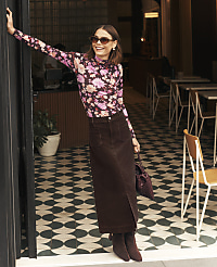 A smiling woman in a floral top and long brown skirt stands in a doorway against a background of a cafe with patterned tile flooring and wooden chairs. carousel Product Image 4