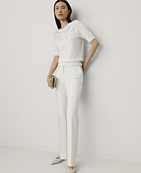 A woman wearing a white embellished short-sleeved top and white trousers stands against a plain white wall. carousel Product Image 1