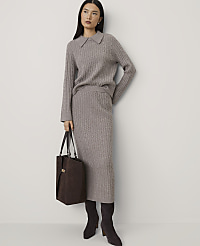 A woman wearing a grey cable-knit sweater and skirt set with dark brown boots and a handbag stands against a plain white wall. carousel Product Image 1