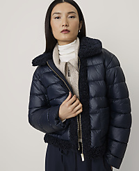 A woman wearing a navy blue puffer jacket over a beige sweater stands against a plain, light-colored background. carousel Product Image 1