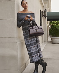 A woman in a grey off-the-shoulder top and plaid skirt leans against a stone wall on a city sidewalk. carousel Product Image 4