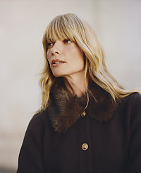 A blonde woman wearing a dark coat with a faux-fur collar stands against a blurred, neutral-toned background. carousel Product Image 4