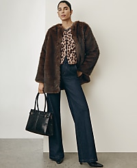 A woman wearing a brown faux-fur coat, leopard print top, and wide-leg jeans stands holding a black handbag against a plain white wall. carousel Product Image 3