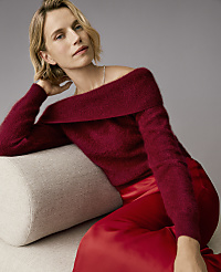 A woman wearing a burgundy off-the-shoulder sweater and red satin trousers leans against a beige bolster pillow against a plain grey background. carousel Product Image 3
