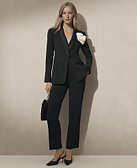A woman wearing a black tailored suit with a white floral accent stands against a plain, neutral-toned wall. carousel Product Image 4