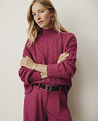 A blonde woman wearing a magenta cable-knit sweater and matching trousers stands with crossed arms against a plain, light-colored wall. carousel Product Image 2