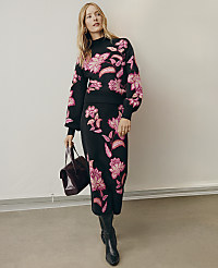 A woman wearing a black floral knit set and black boots stands holding a dark purple handbag against a plain white wall and grey floor. carousel Product Image 4