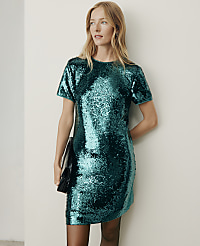 A woman wearing a shimmering teal sequin dress and holding a black clutch stands against a plain, light-colored wall. carousel Product Image 3