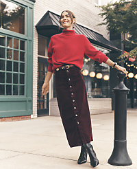 A smiling woman in a red sweater and long burgundy skirt leans against a black bollard on a city sidewalk in front of storefronts. carousel Product Image 2
