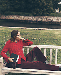 A woman in a red cardigan and burgundy trousers sits on a wooden bench holding a book, with a grassy lawn and a stone riverbank in the background. carousel Product Image 4