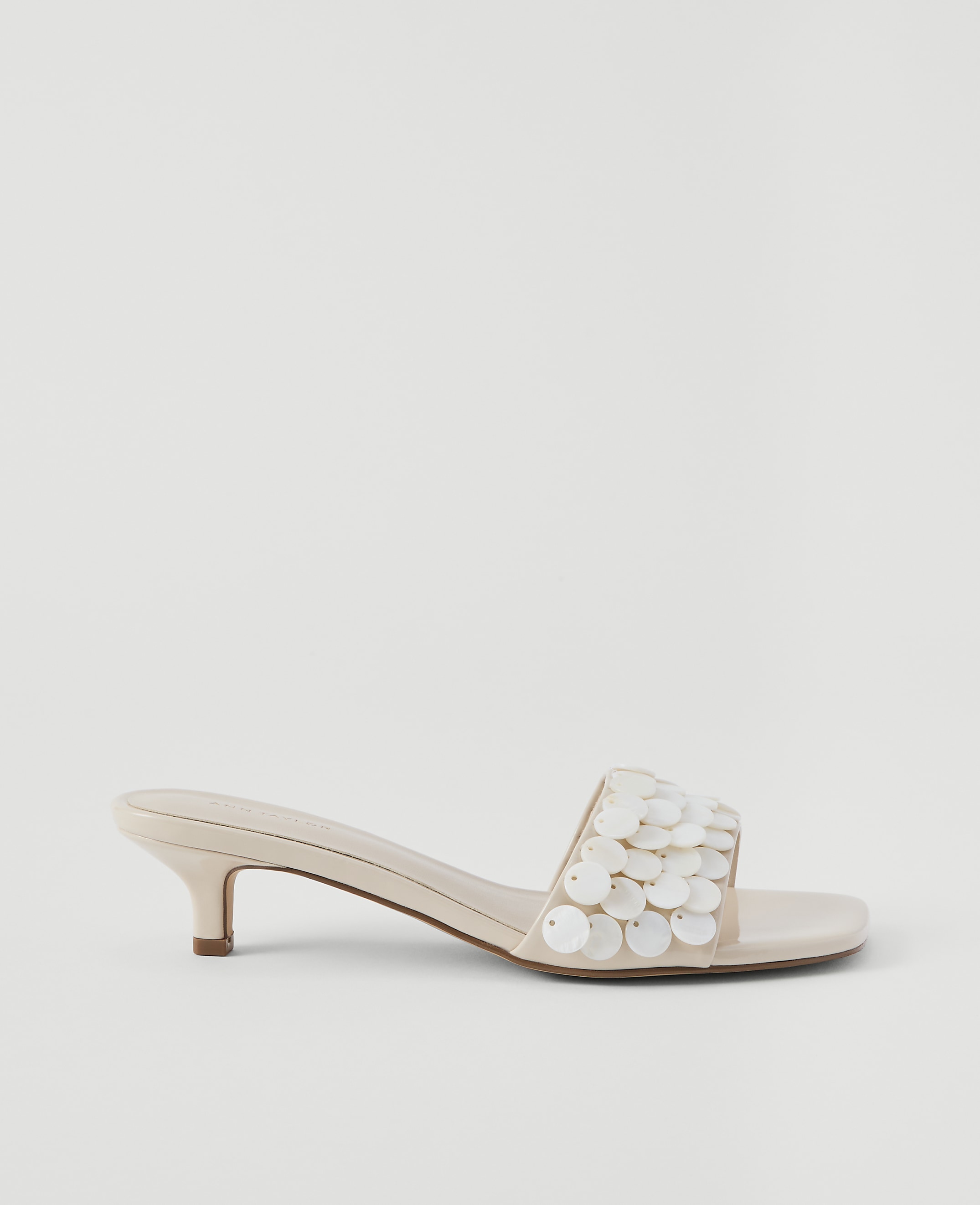 A cream-colored kitten heel sandal with a sequined strap rests on a plain, light gray background.