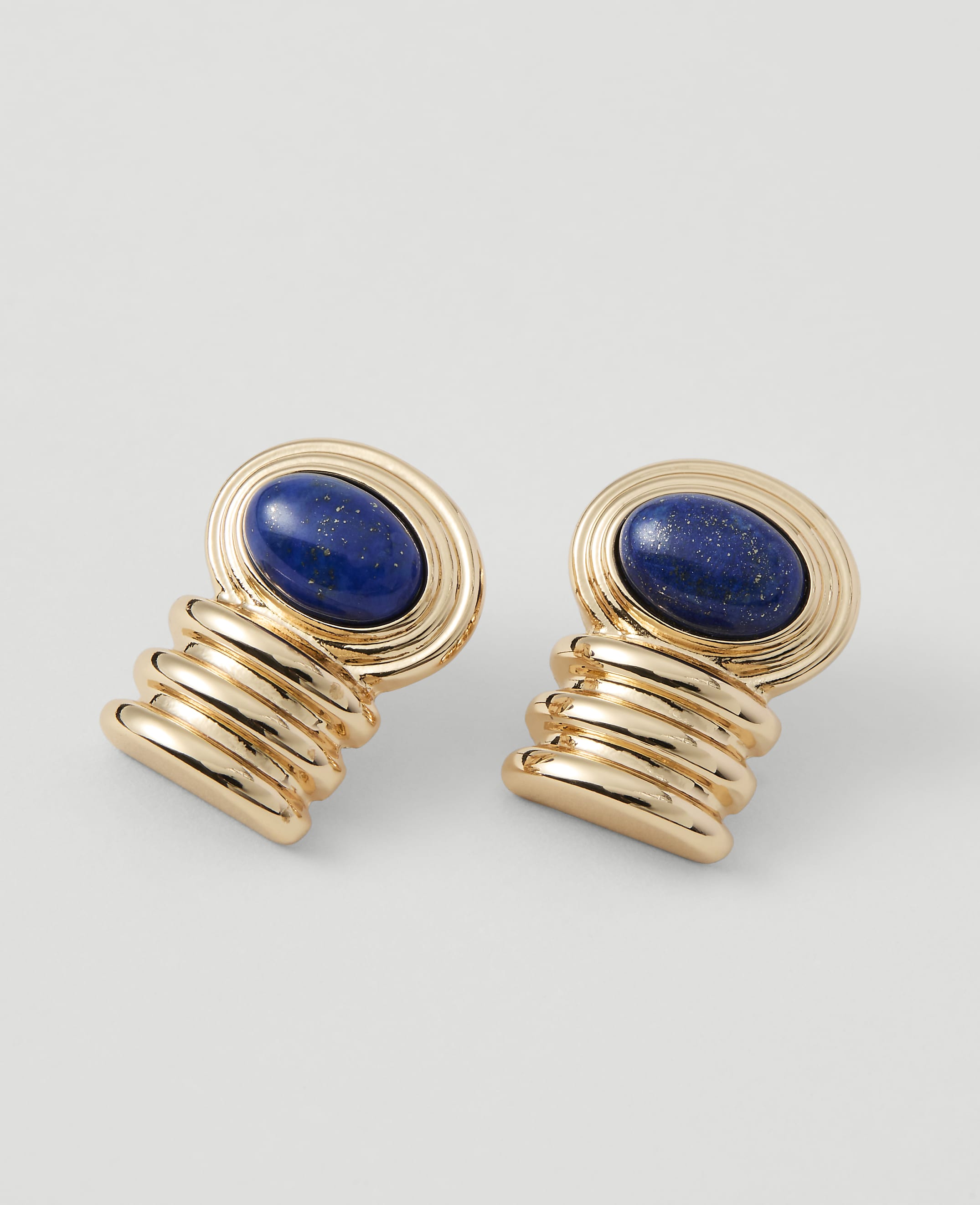 A pair of gold earrings with oval lapis lazuli stones are displayed against a plain, light gray background.