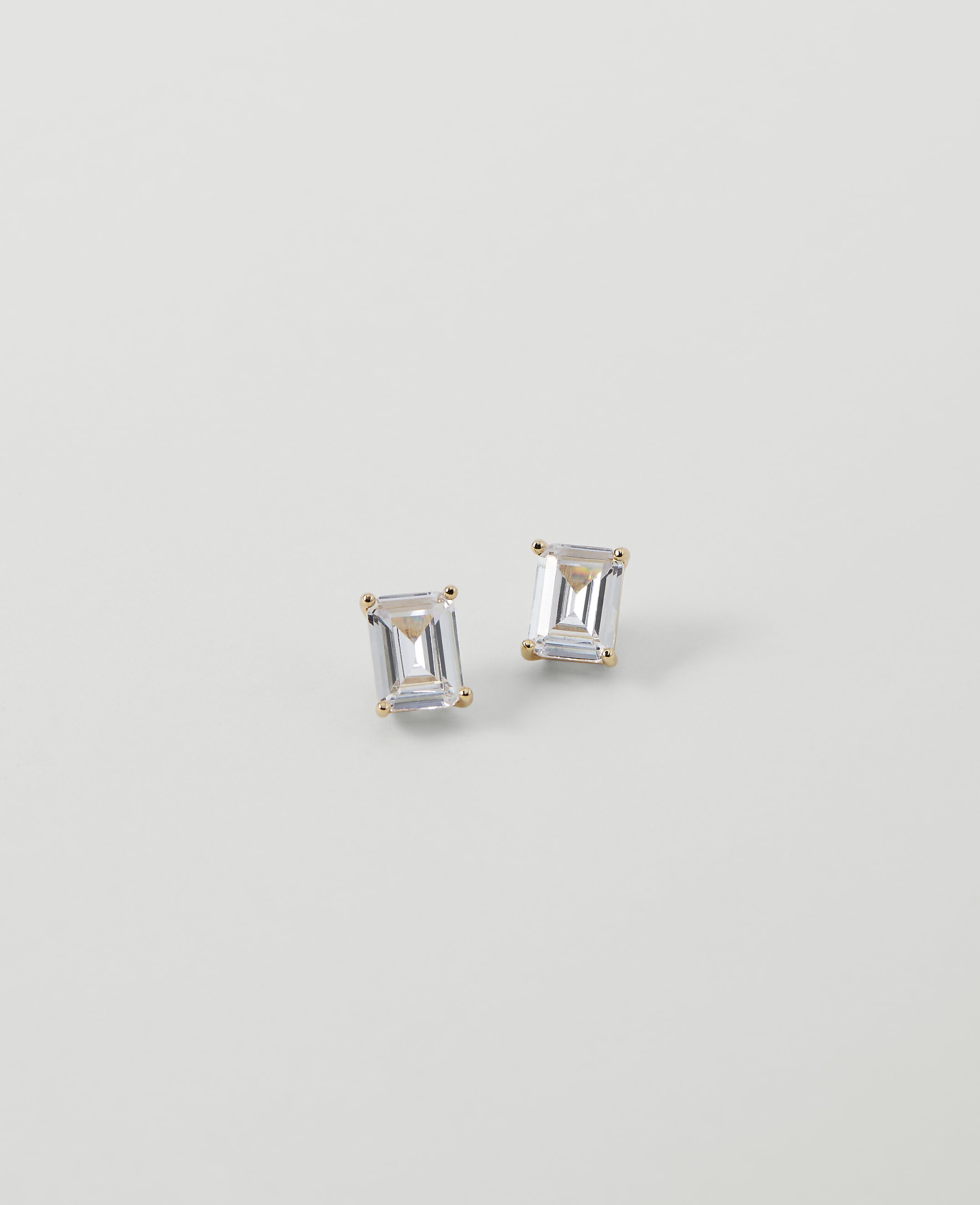Two emerald-cut diamond stud earrings rest on a plain, light gray background.