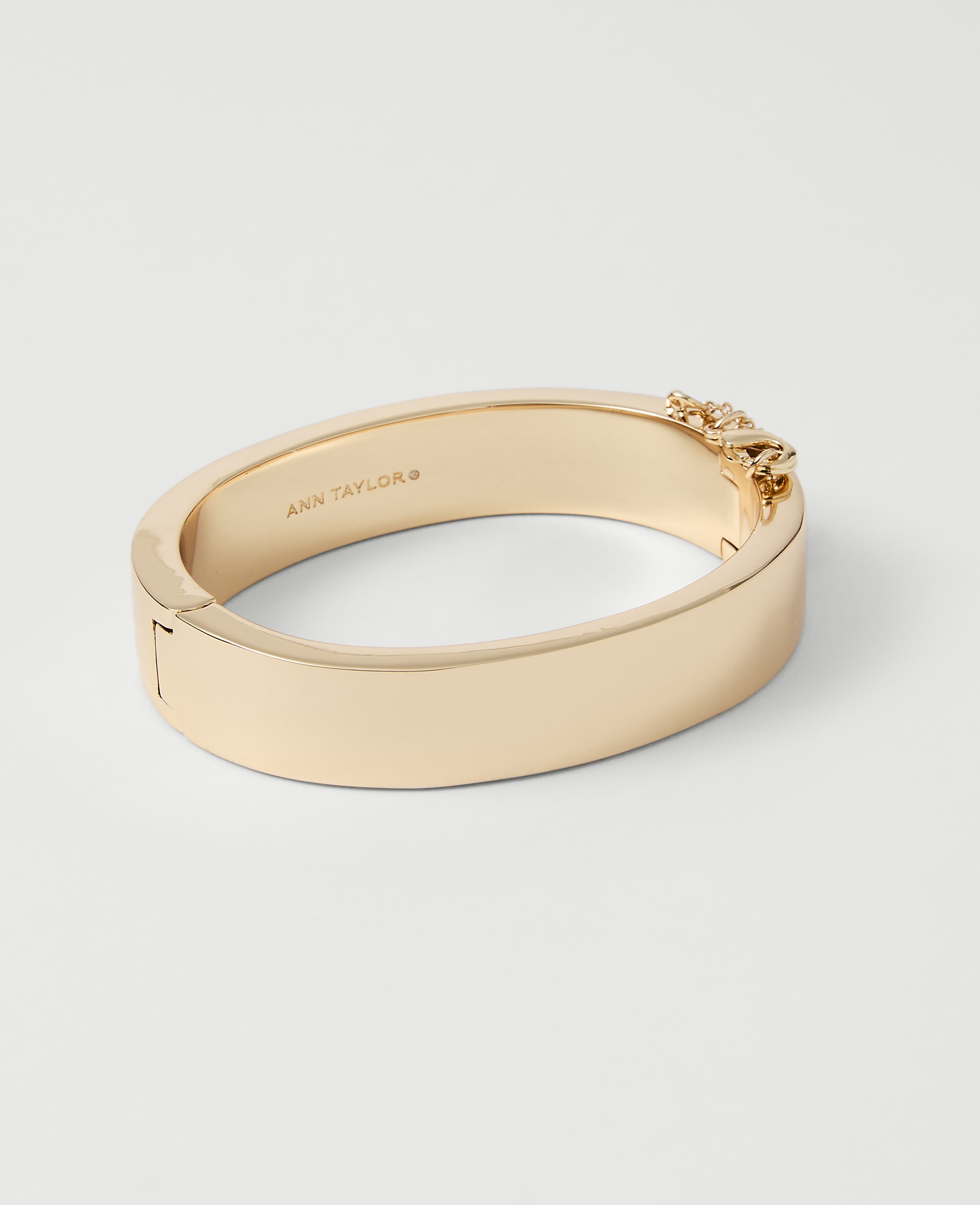 A gold bangle bracelet with a chain detail on the clasp rests on a plain white background.