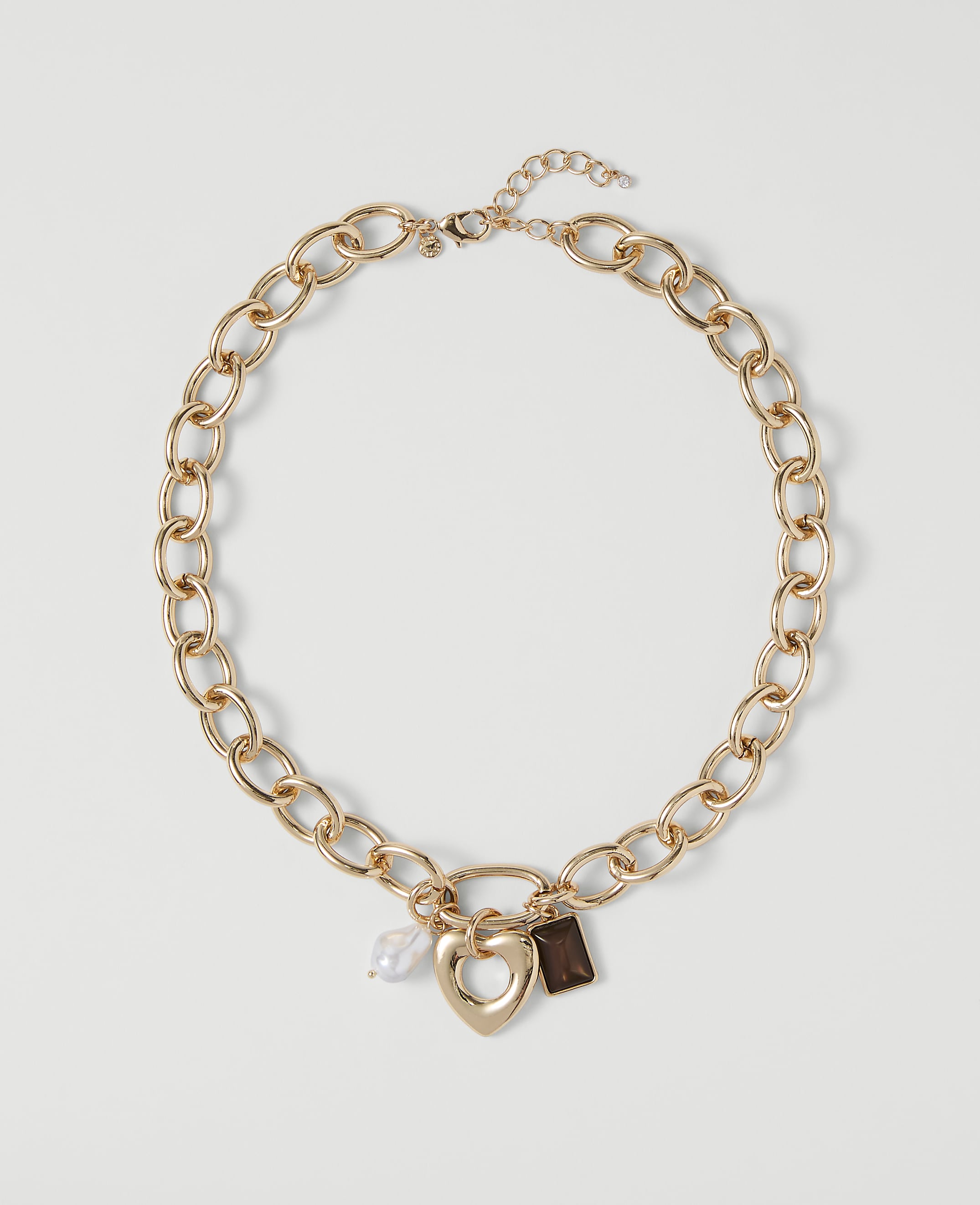 A gold chain necklace with a heart pendant and two charms rests on a plain white background.