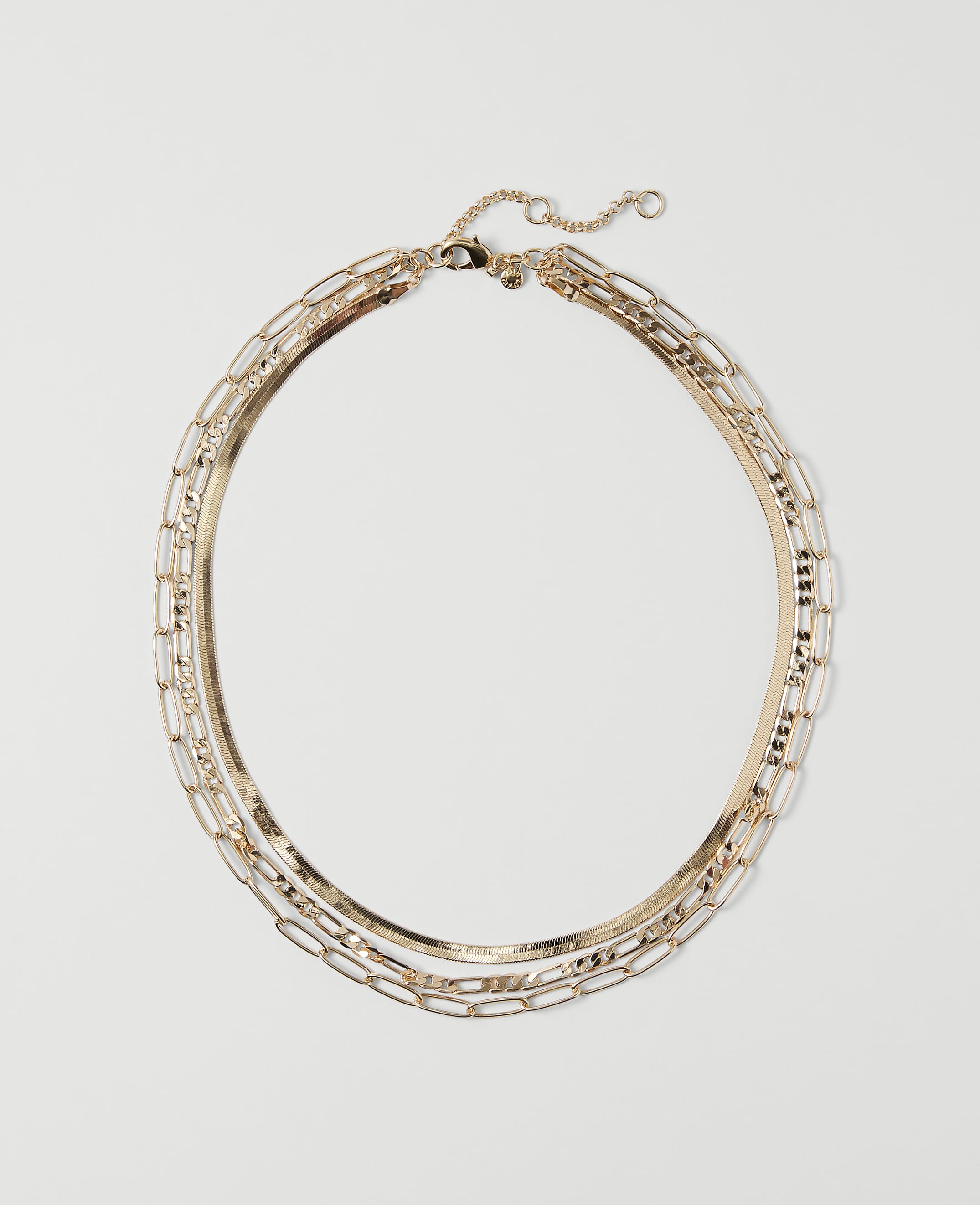 A layered gold necklace with a herringbone chain and rectangular links rests on a plain white background.