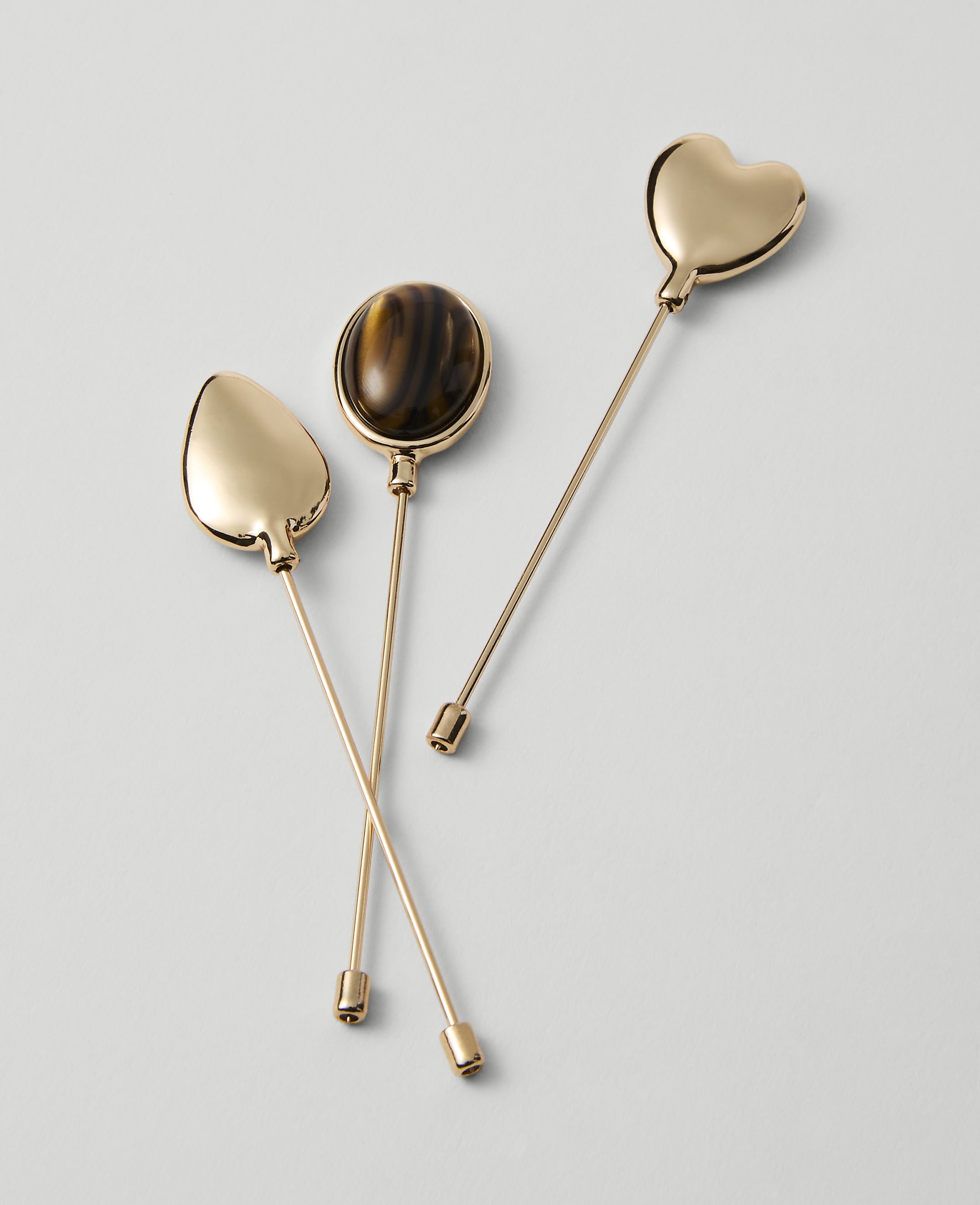 Three gold lapel pins, one teardrop-shaped, one oval with a tiger's eye stone, and one heart-shaped, rest on a plain white background.