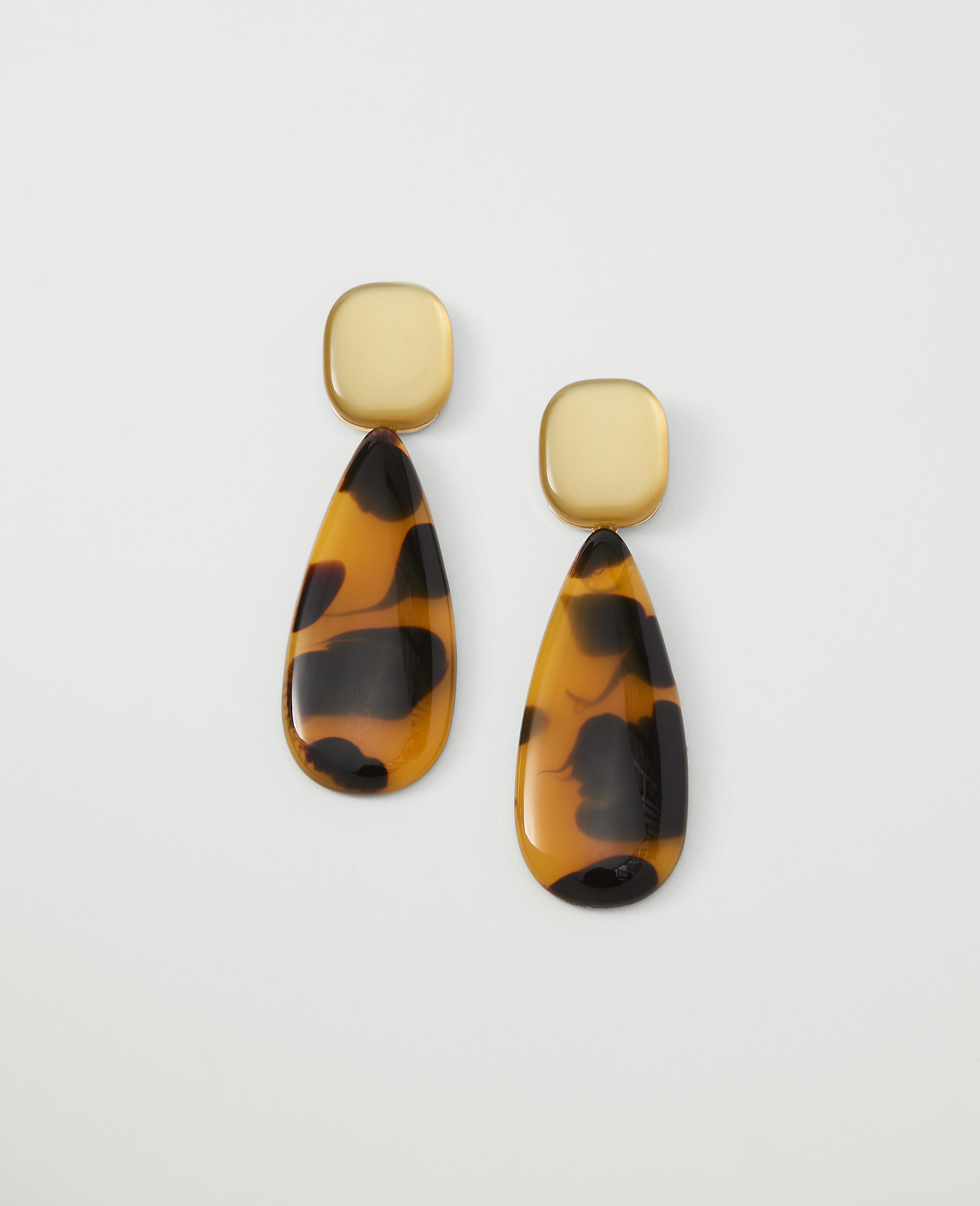 A pair of tortoise-shell teardrop earrings with gold square tops hang against a plain white background.