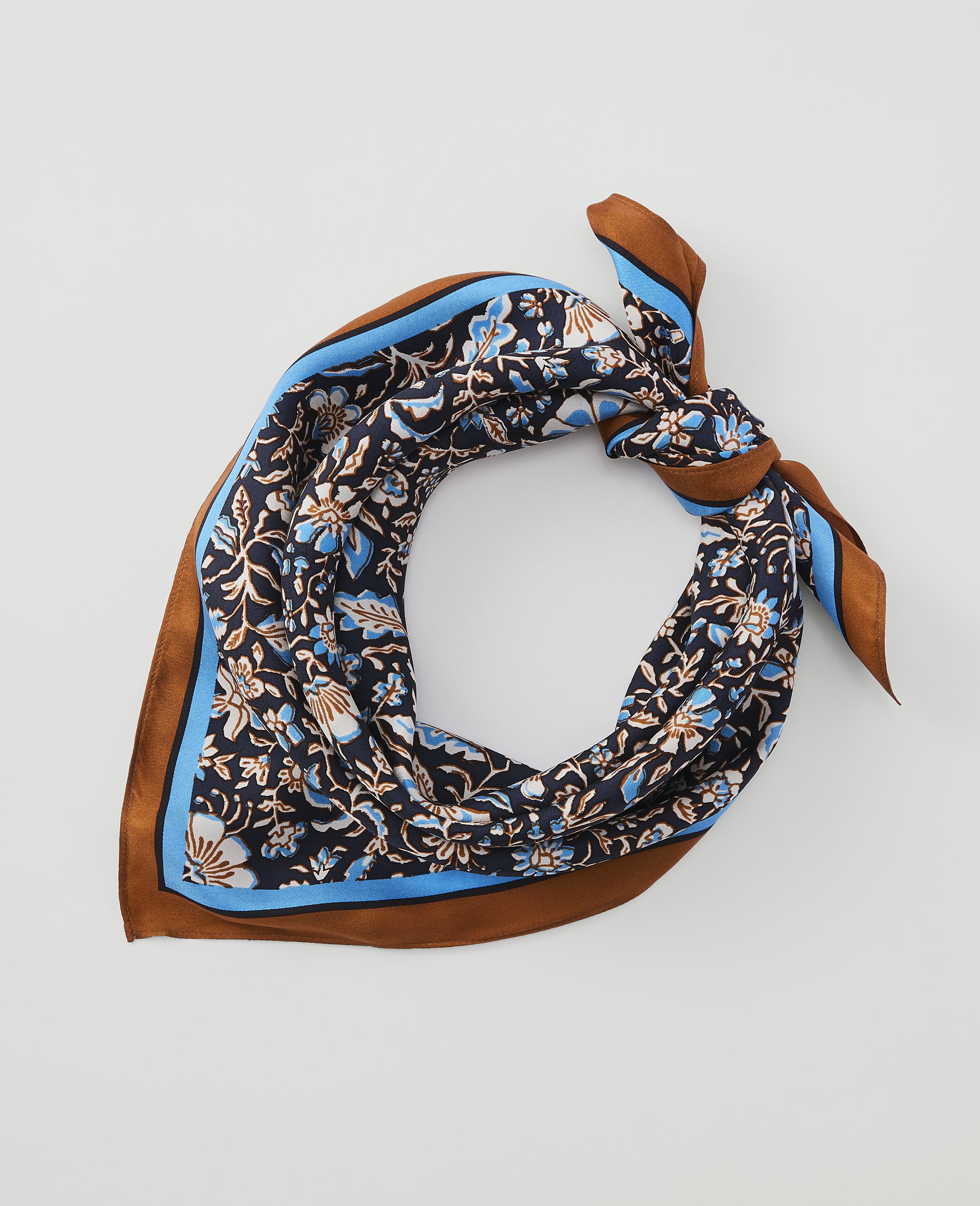 A dark blue floral patterned silk scarf with brown and light blue trim is tied in a knot against a plain white background.