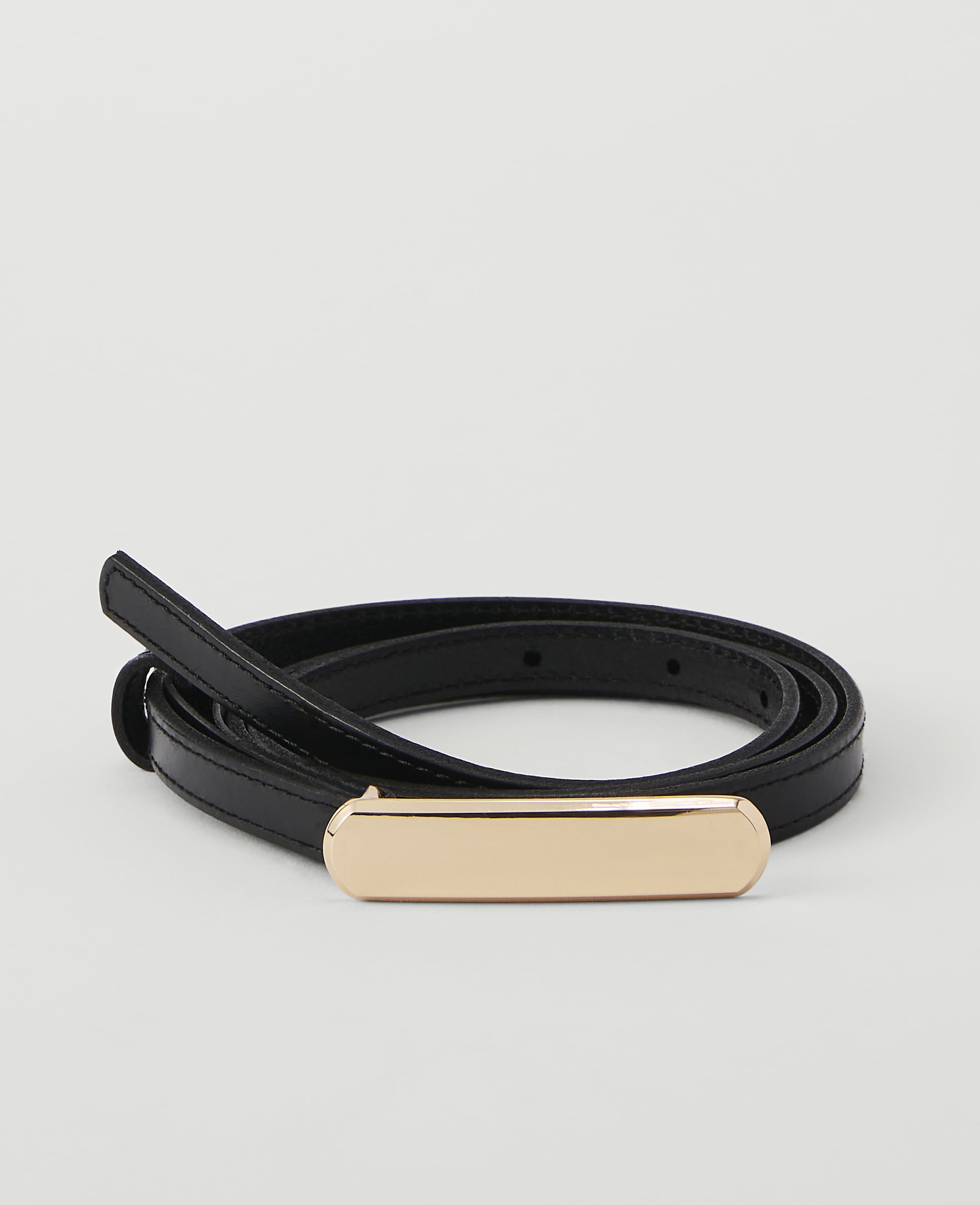 A thin black belt with a gold buckle is coiled on a plain white background.