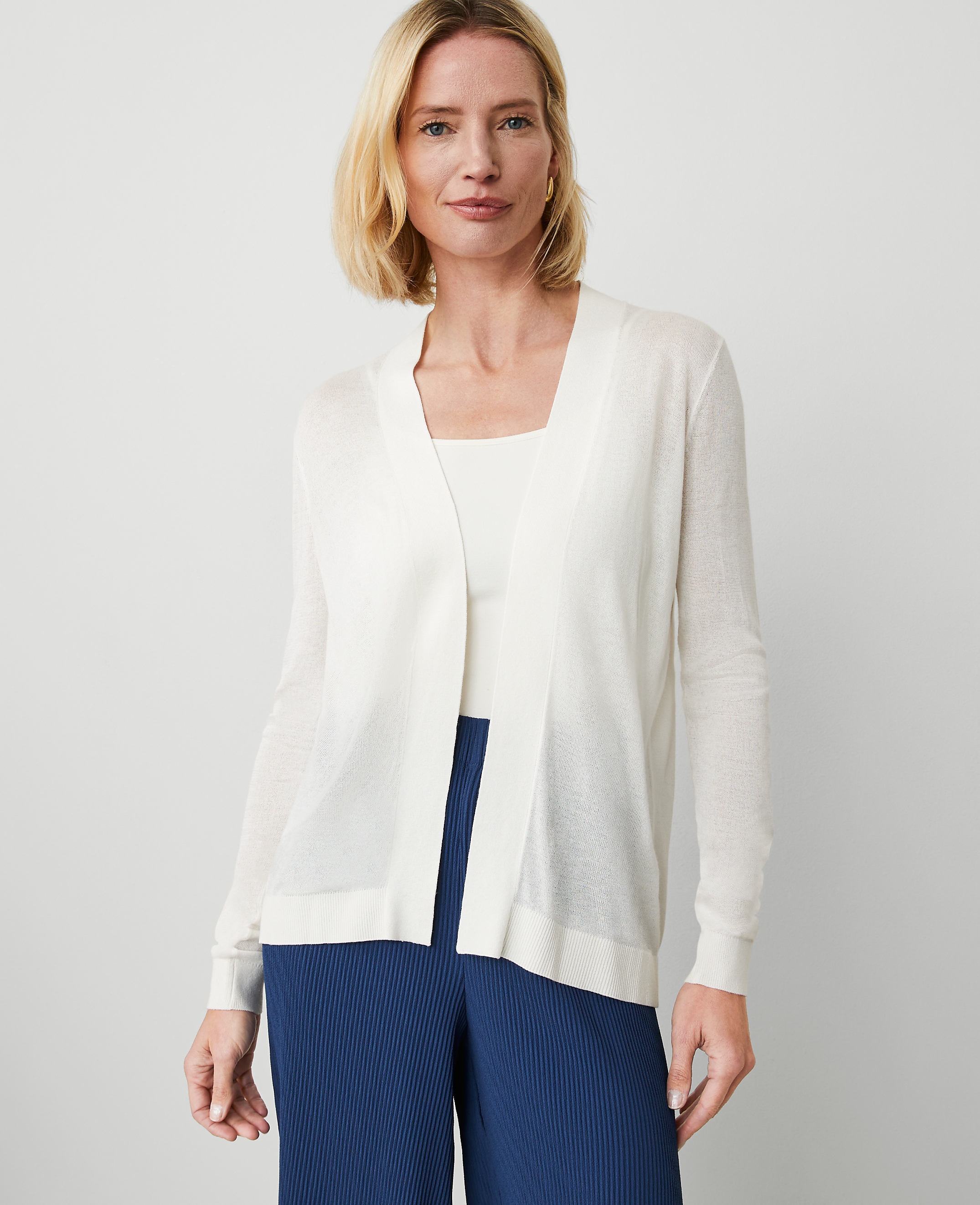 A woman in a white cardigan and blue pants stands against a plain white background.