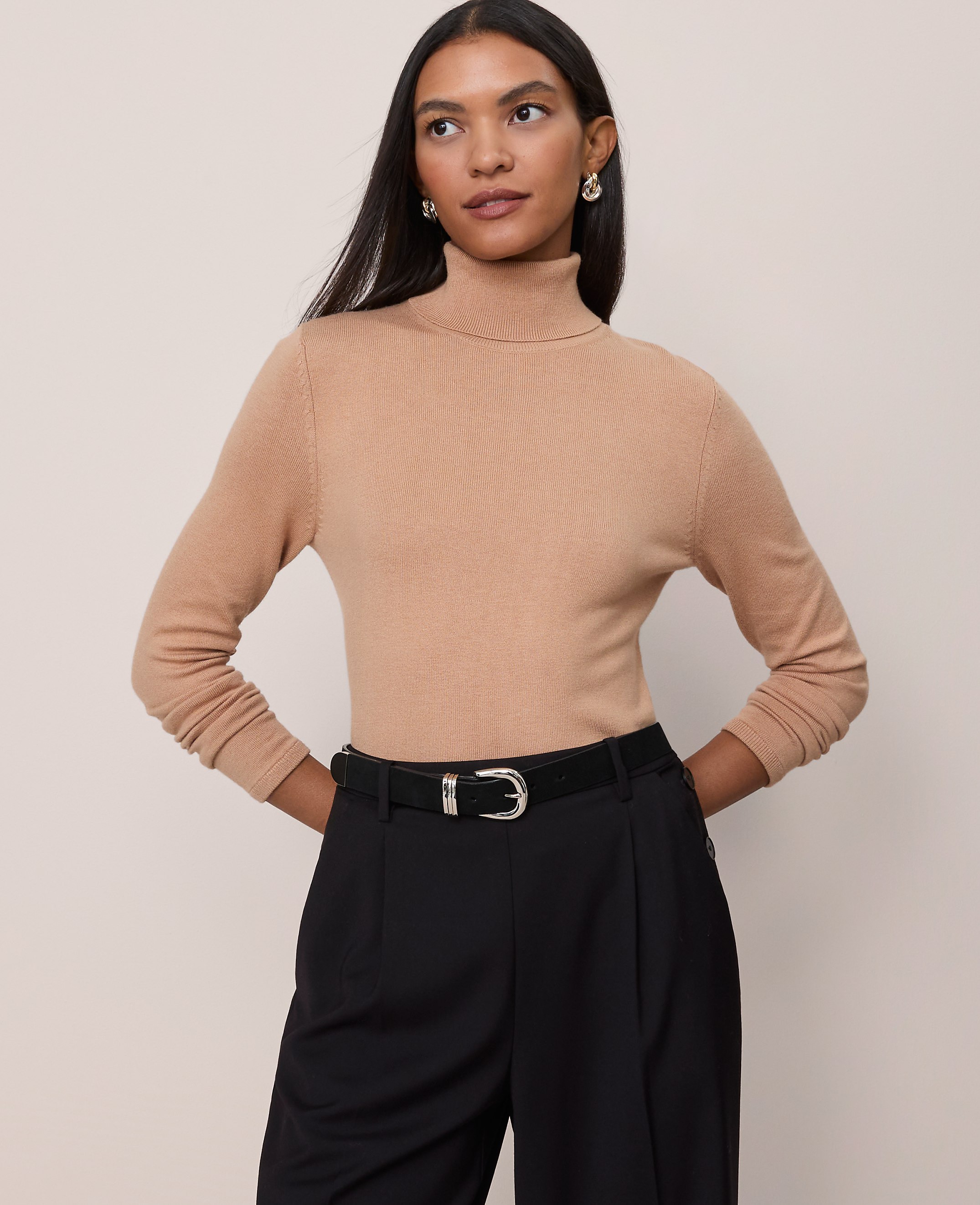 A woman in a tan turtleneck and black pants stands against a plain beige background.