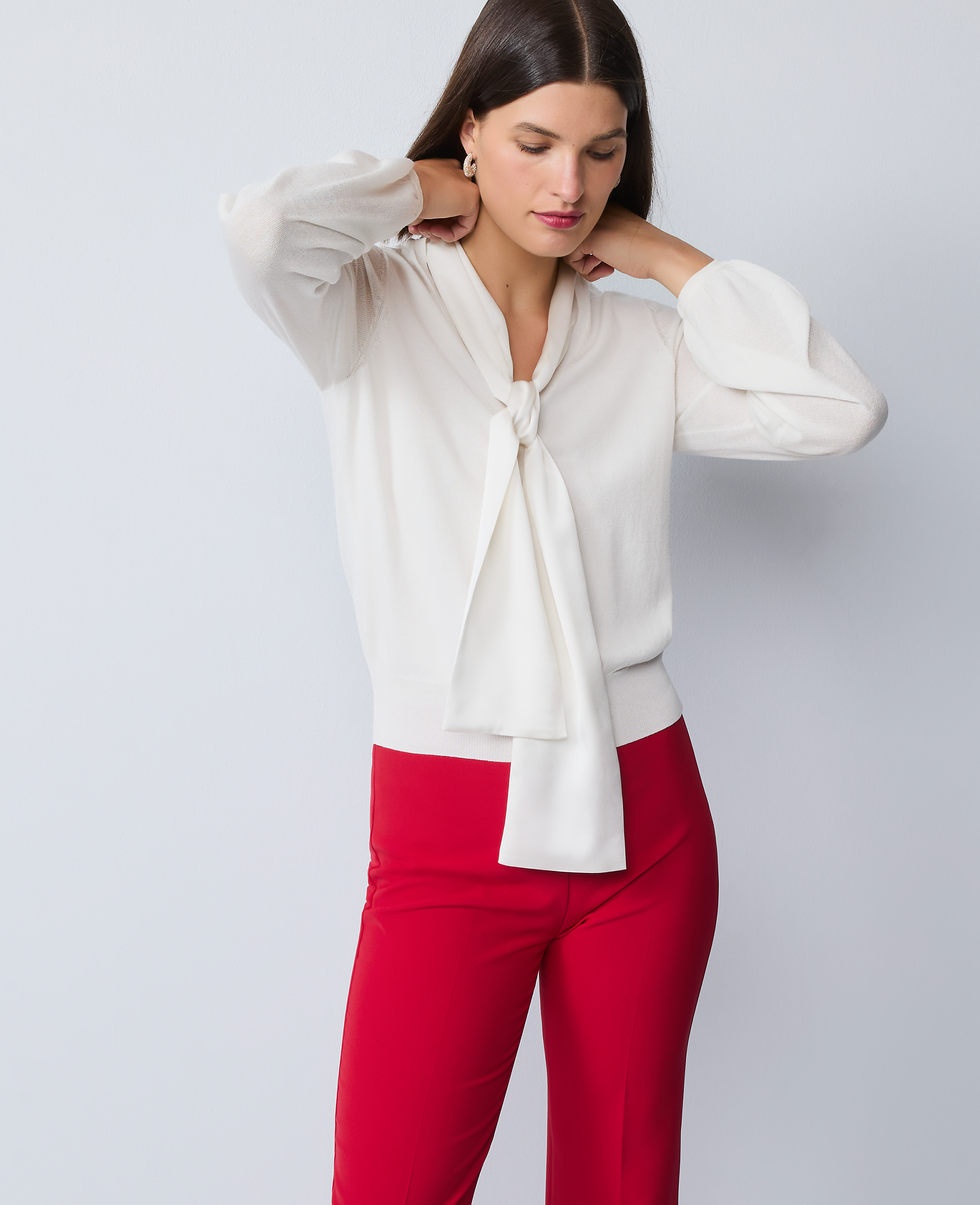 A woman in a cream bow-neck blouse and red pants stands against a plain gray background.