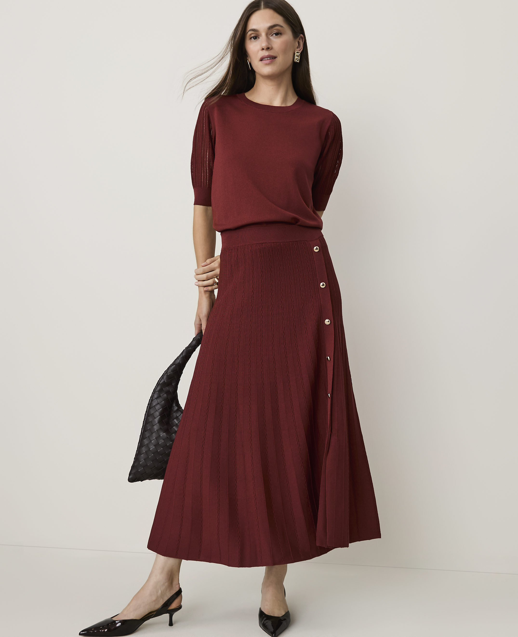 A woman in a burgundy knit set and black heels stands against a plain white background, holding a black woven bag.