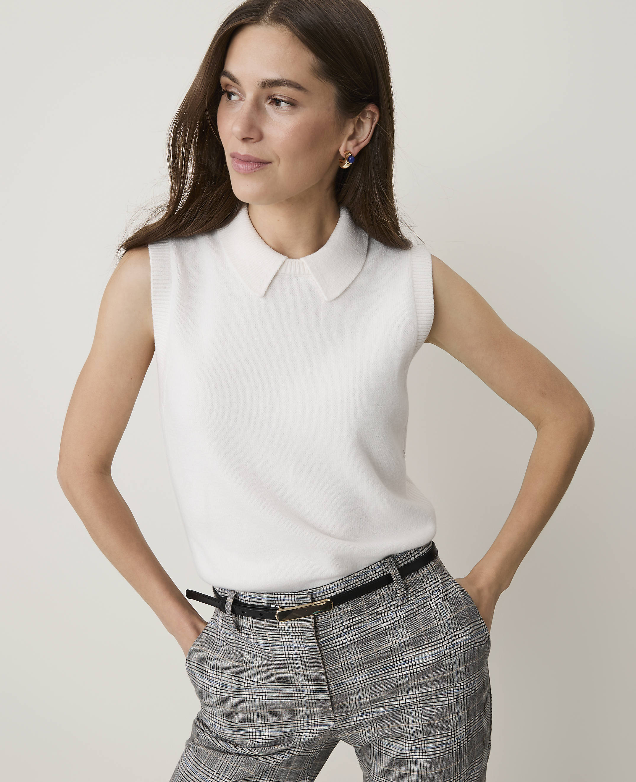 A woman in a white sleeveless collared top and plaid pants stands against a plain white background.