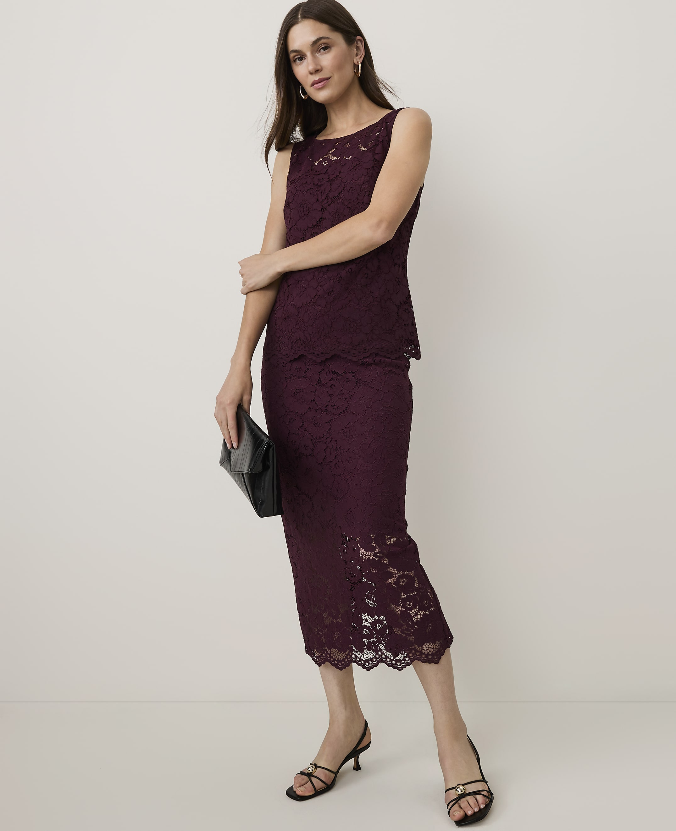 A woman in a burgundy lace dress and black heels stands with a clutch in front of a plain background.