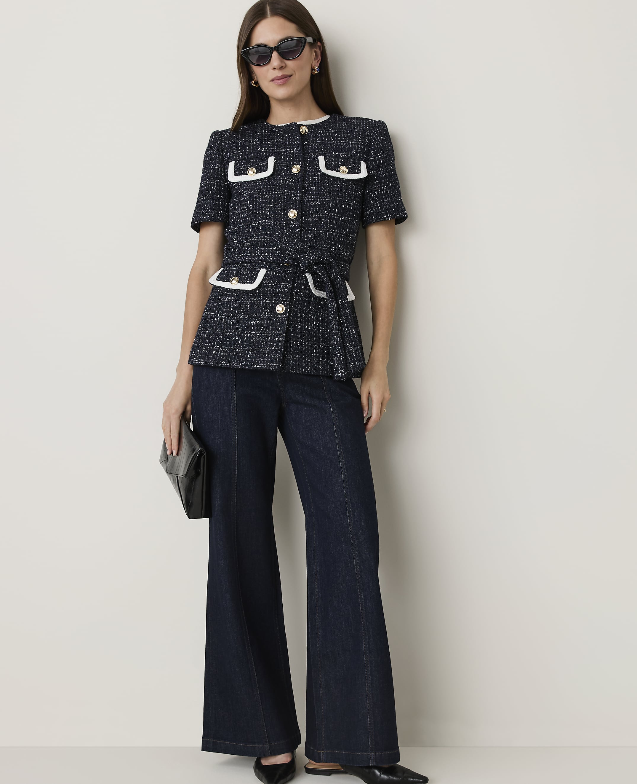 A woman in a navy tweed jacket and dark flared jeans stands against a plain, light-colored wall, holding a black clutch.