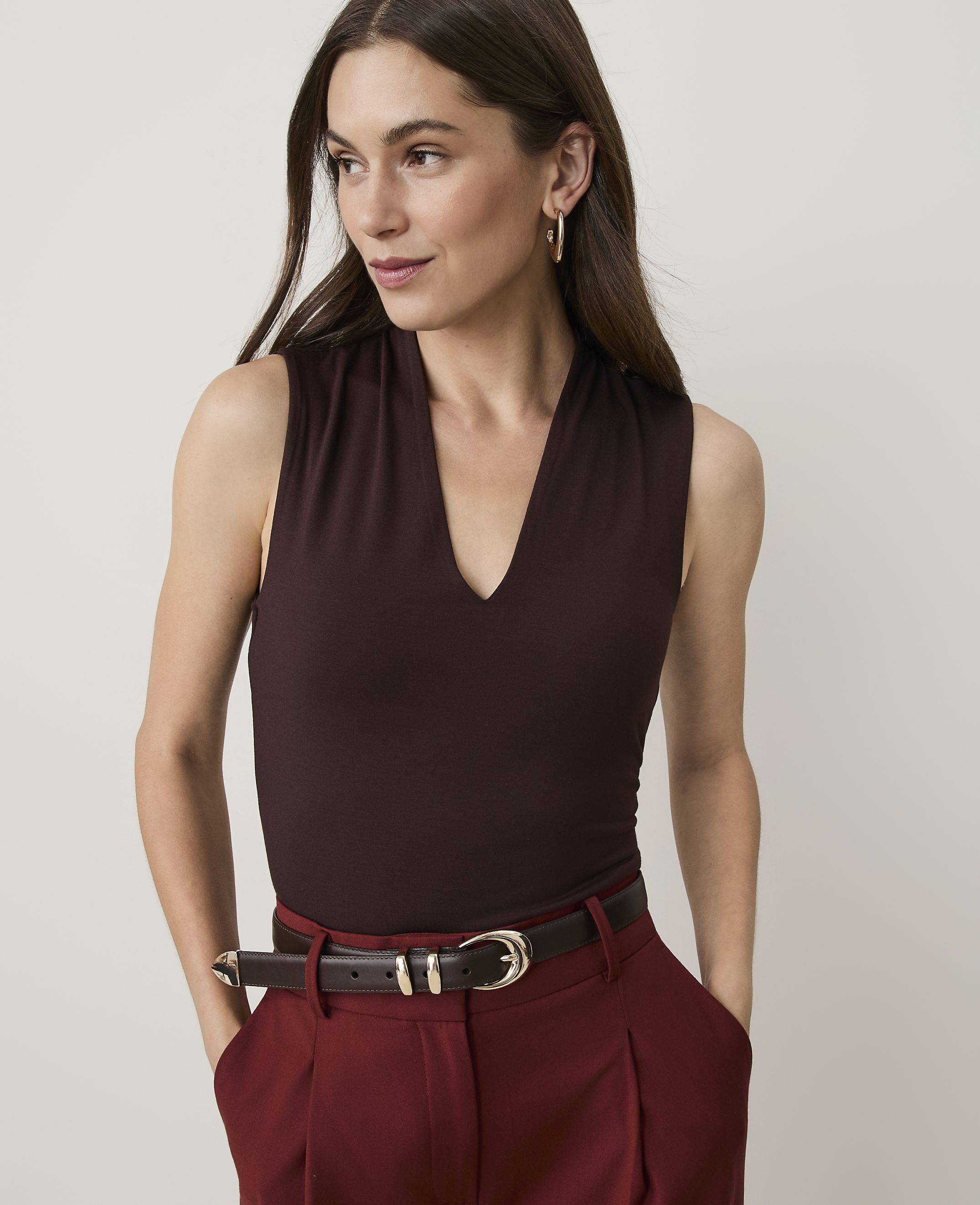 A woman in a dark brown V-neck top and maroon pants stands against a plain background.