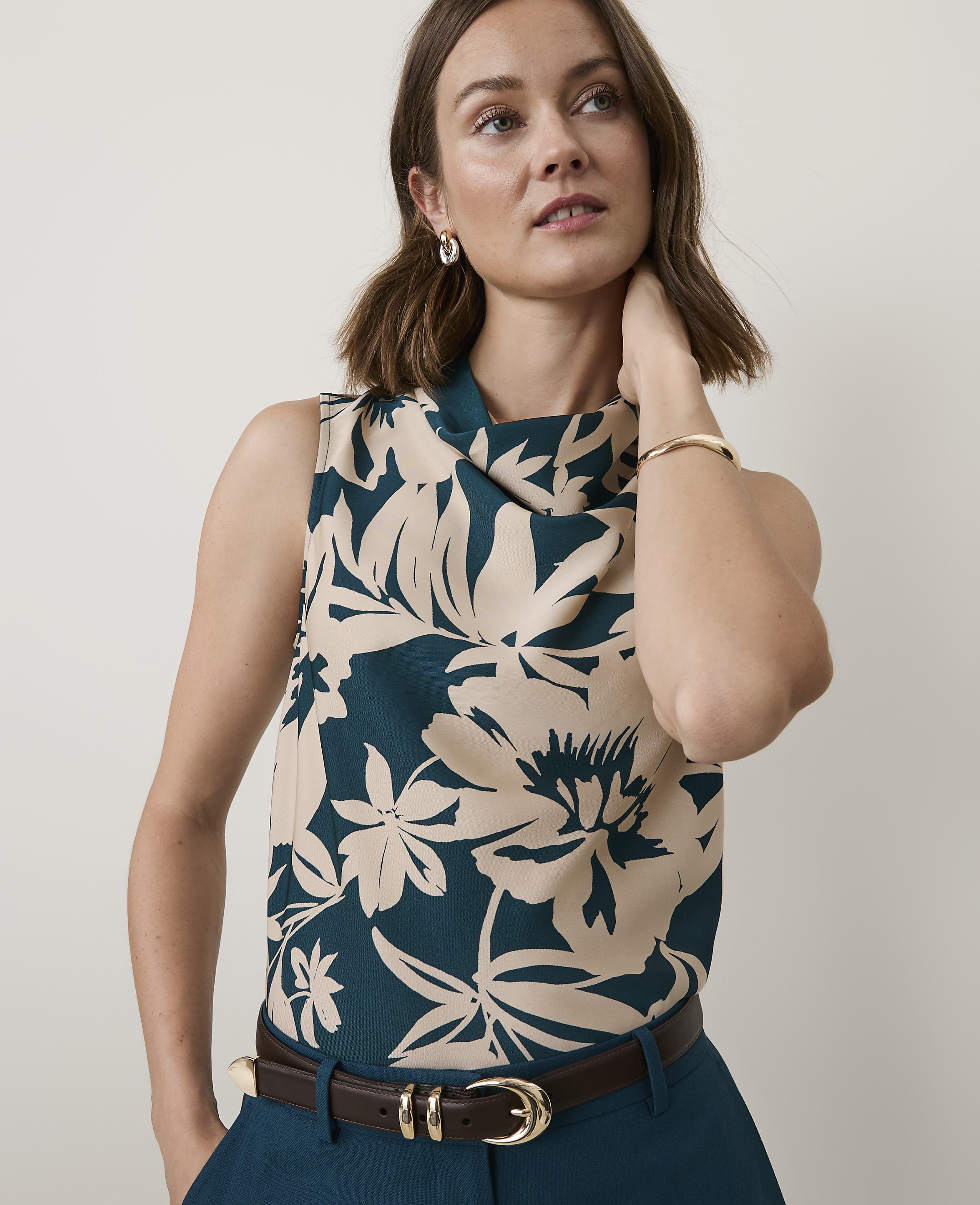 A woman in a floral top and dark pants stands against a plain, light background.