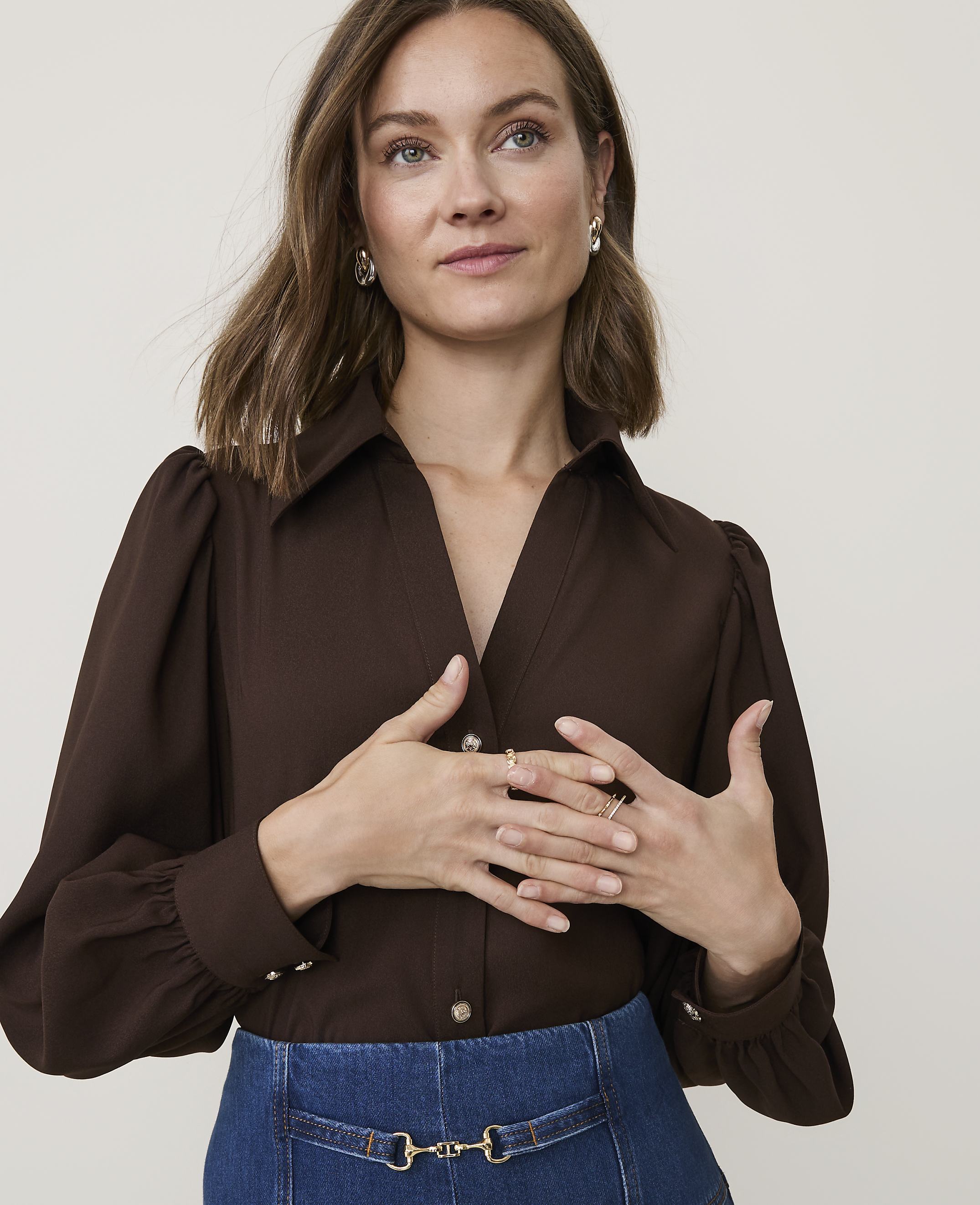 A woman in a brown blouse and blue skirt poses with her hands near her chest against a plain background.