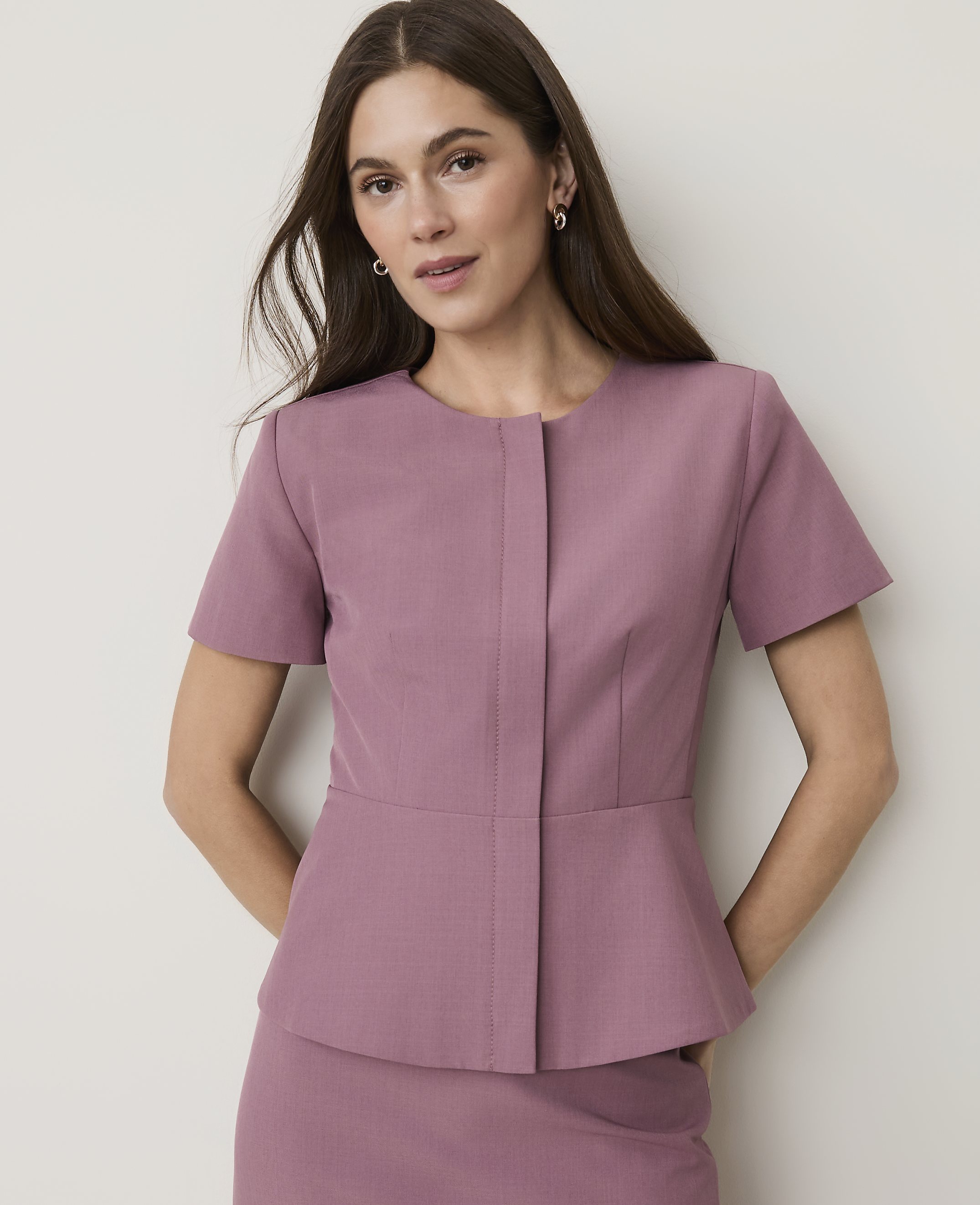 A woman in a mauve peplum jacket and skirt stands against a plain, light-colored background.
