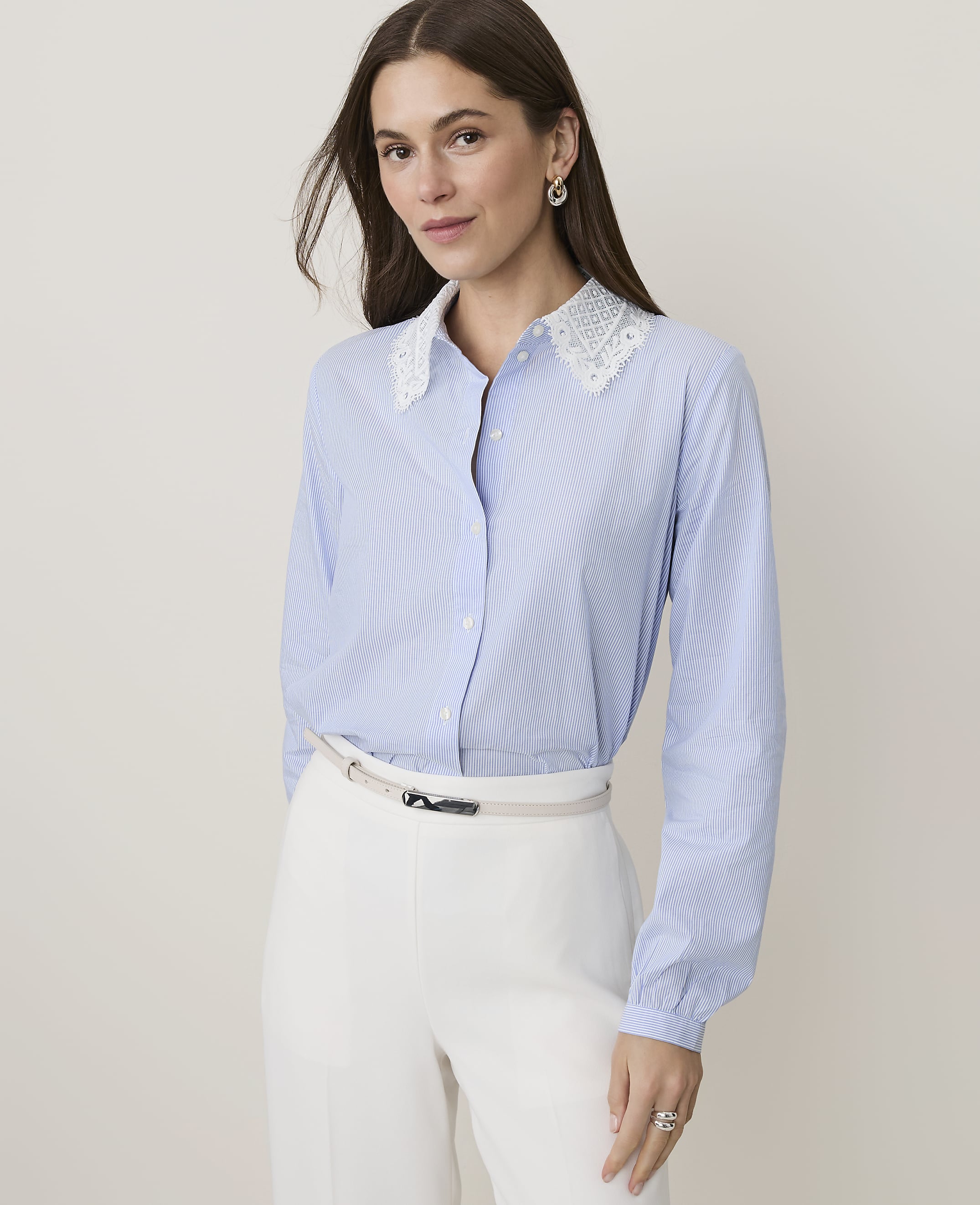 A woman in a blue pinstripe shirt with a lace collar and white pants stands against a plain, light background.