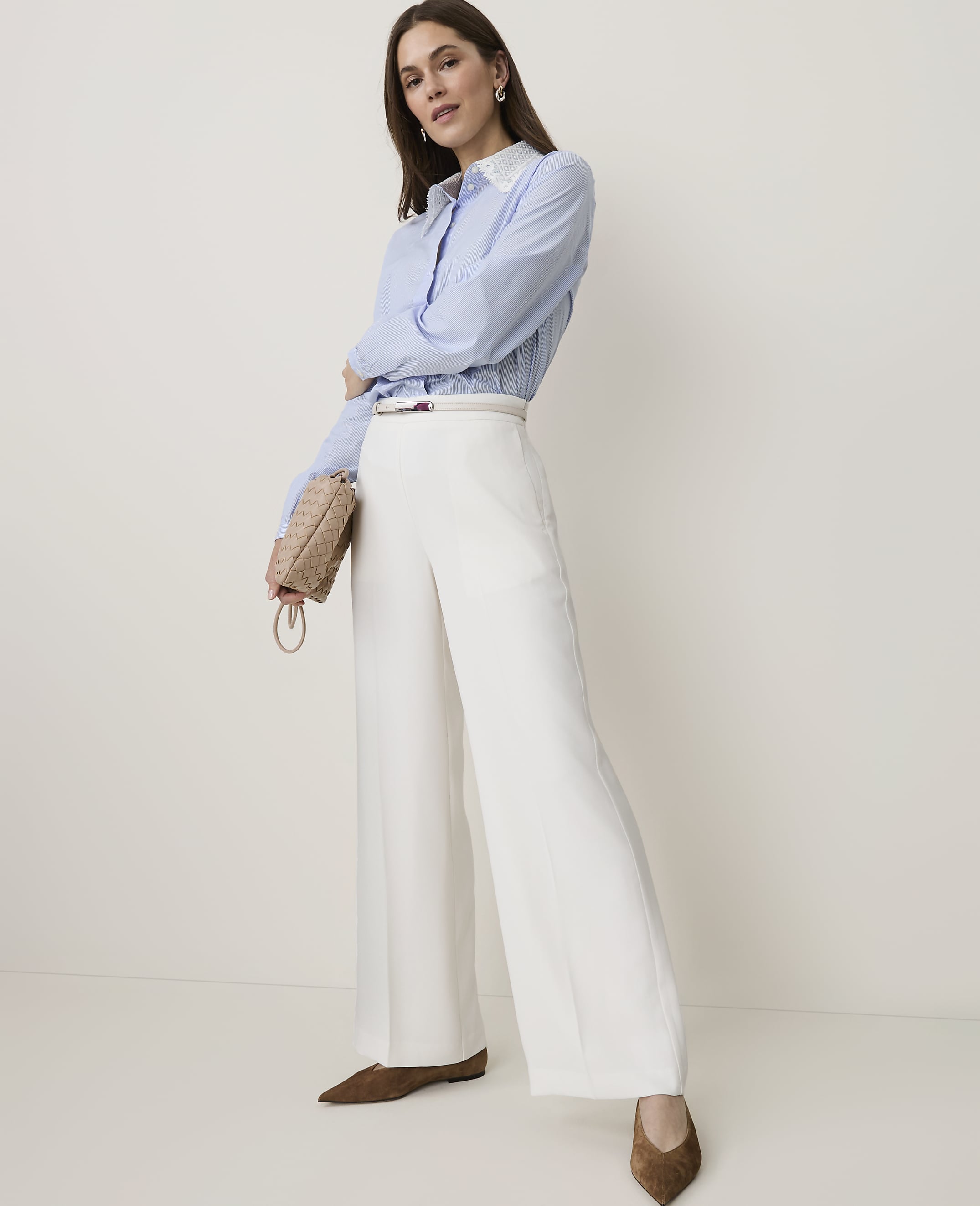 A woman in a blue striped shirt and white wide-leg pants stands against a plain, light-colored background, holding a small woven purse.