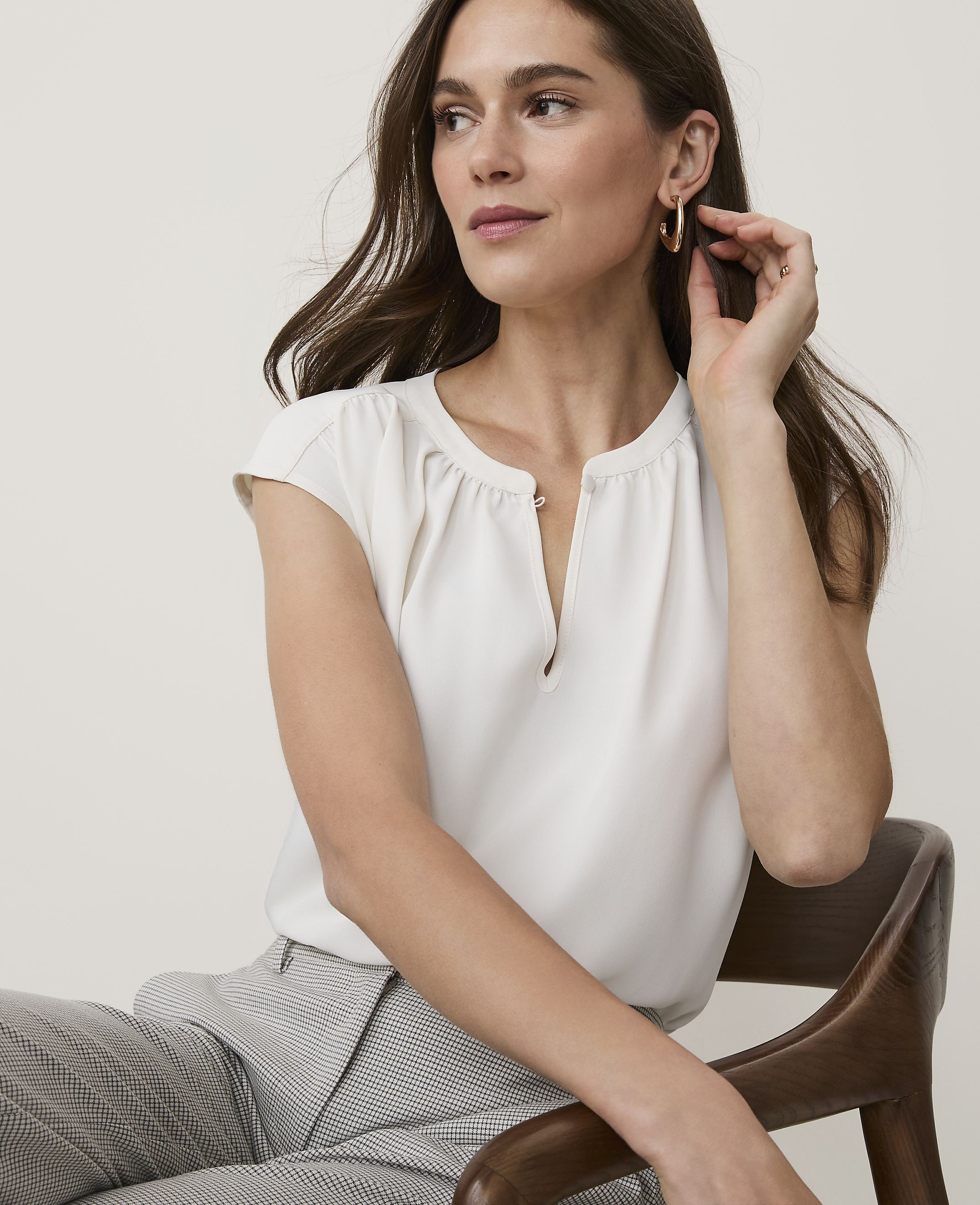 A woman in a cream blouse and patterned pants sits on a wooden chair against a plain background.