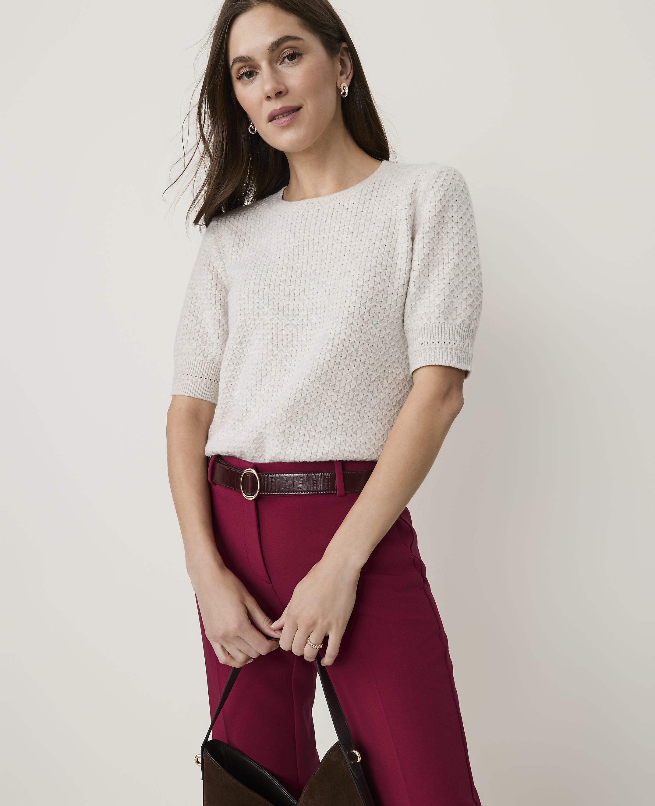 A woman in a light-colored textured sweater and maroon pants holds a dark purse against a plain, light background.