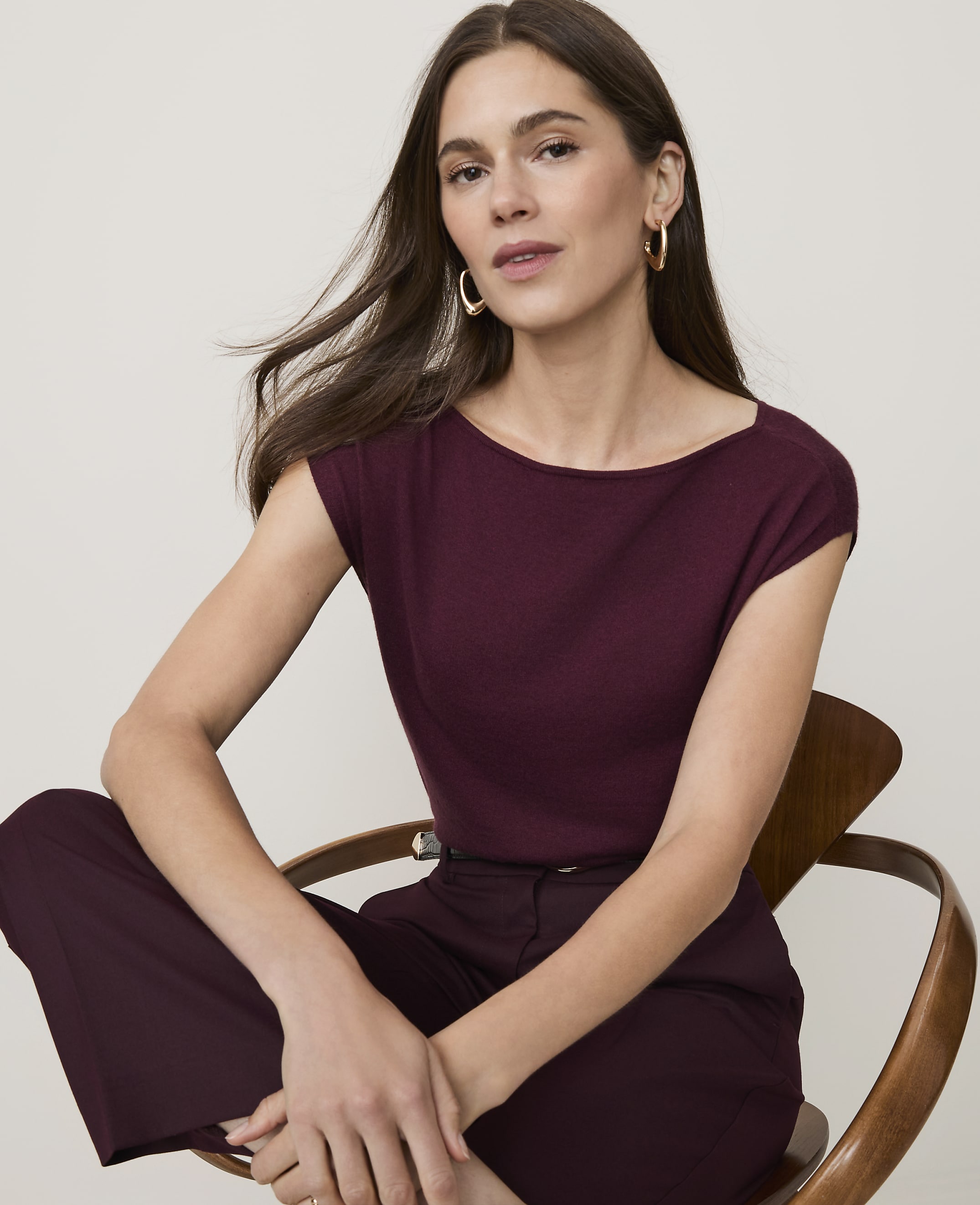 A woman in a maroon top and pants sits on a wooden chair against a plain, light background.