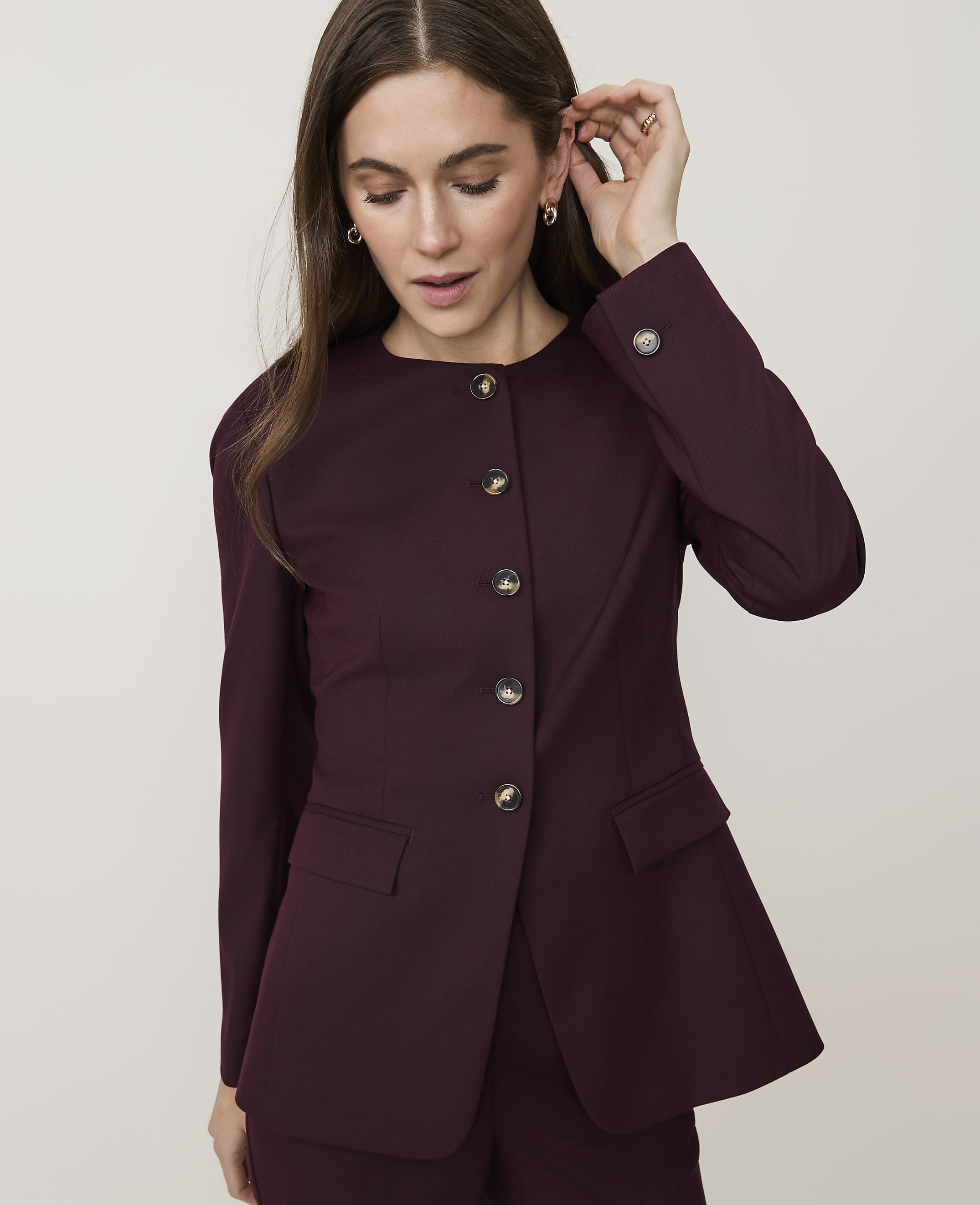 A woman in a burgundy blazer with round buttons adjusts her earring against a plain white background.